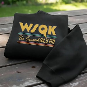 Peut inclure: Un sweat-shirt noir avec l'inscription "WSQK The Squawk 94.5 FM" en or, turquoise, orange et violet. Le sweat-shirt est plié sur une surface en bois, avec une manche visible. Le design est de style rétro.