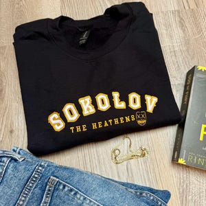 K&ouml;nnte beinhalten: Schwarzes Sweatshirt mit gelb-wei&szlig;en Buchstaben "SOKOLOV" und "THE HEATHENS" darunter. Eine goldene Kette, ein Buch und blaue Jeans sind ebenfalls im Bild.