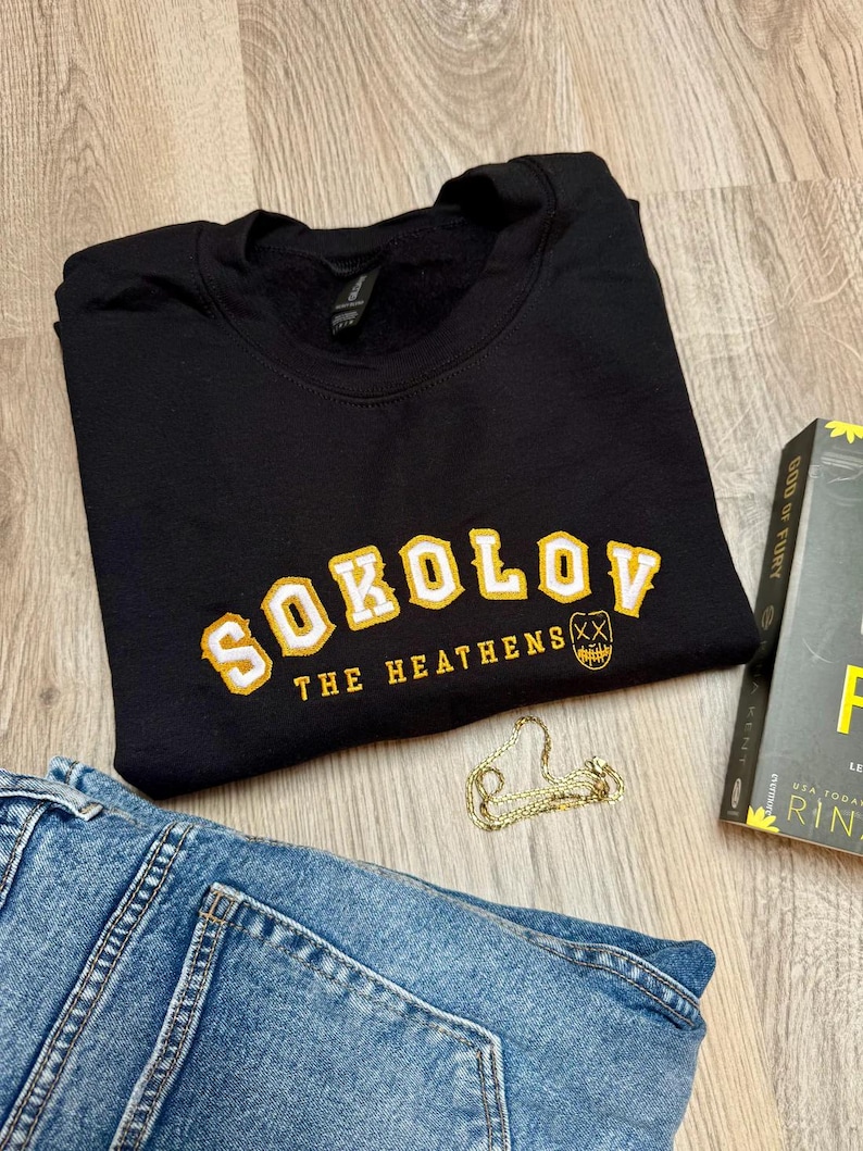 K&ouml;nnte beinhalten: Schwarzes Sweatshirt mit gelb-wei&szlig;en Buchstaben "SOKOLOV" und "THE HEATHENS" darunter. Eine goldene Kette, ein Buch und blaue Jeans sind ebenfalls im Bild.