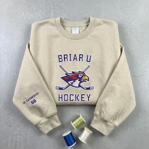 May include: A tan crewneck sweatshirt with the text "Briar U Hockey" and a logo of a hawk with crossed hockey sticks. The left sleeve has the text "Di Laurentis 66" in blue. Three spools of thread are at the bottom.