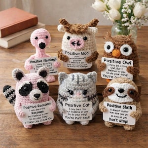 Peut inclure: Ensemble de six figurines d'animaux crochetés, dont un flamant rose, une vache, un hibou, un raton laveur, un chat et un paresseux. Chacun tient une pancarte avec un message inspirant. Les figurines sont de couleurs variées.