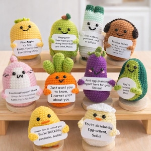 Peut inclure: Une collection de peluches de fruits et légumes crochetés à la main, chacune avec un visage souriant et une petite pancarte. Les peluches comprennent une pomme, une poire, une échalote, une pomme de terre, un requin, une carotte, une aubergine, un avocat, un canard et un œuf.
