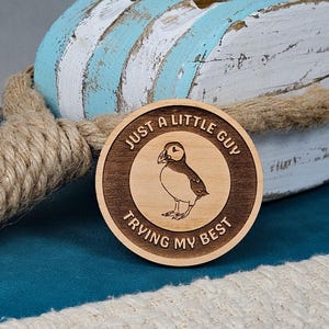 May include: A round wooden coaster with a laser-etched design featuring a puffin illustration and the text "JUST A LITTLE GUY TRYING MY BEST". The coaster is set against a backdrop of a distressed, light blue and white wooden piece and rope.