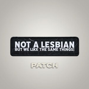 May include: A rectangular black embroidered patch with a white border and white text that reads NOT A LESBIAN BUT WE LIKE THE SAME THINGS! The background of the patch features a subtle, dark gray topographic map pattern across the fabric surface.