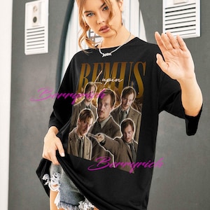 May include: Black t-shirt featuring a graphic of Remus Lupin from the Harry Potter series. The design includes the name "Remus Lupin" in gold lettering above a collage of images of the character. The shirt is worn by a person.
