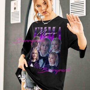 May include: Black t-shirt with a purple and white design featuring the name "EMMA Myers" and a collage of images. The shirt has a crew neck and three-quarter sleeves. The model is wearing denim shorts.