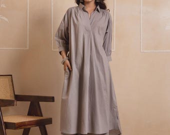 May include: A light gray tunic dress and matching pants. The dress has a collared neckline and long sleeves. The pants are straight-legged. The outfit is worn with sandals. A wooden chair and a round rug are in the background.