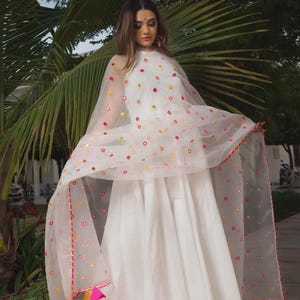 A white dress with a flowing skirt and a sheer, dotted shawl. The shawl is adorned with colorful circular accents and a pink trim. The outfit is completed with gold sandals and colorful tassels.