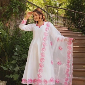 Könnte beinhalten: Weißes Langarmkleid mit rosa Blumenstickerei an den Rändern und am Ausschnitt. Das Kleid wird mit einer passenden Hose und einem transparenten Schal kombiniert, der ebenfalls mit rosa Blumendesigns verziert ist. Das Outfit wird im Freien getragen.
