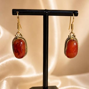 Könnte beinhalten: Ein Paar Ohrhänger mit ovalen, tief orange-roten Steinen in goldfarbenen Metallfassungen mit gedrehtem Rand. Die Ohrringe hängen an goldfarbenen Fischhaken-Verschlüssen und sind an einem schwarzen, T-förmigen Schmuckständer befestigt.