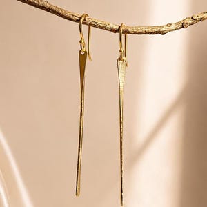 Könnte beinhalten: Ein Paar lange, schlanke goldene Ohrhänger mit einer texturierten, gehämmerten Oberfläche. Jeder Ohrring besteht aus einem dünnen, vertikalen Metallstab, der an einem einfachen Haken befestigt ist und eine helle, reflektierende gelbe Oberfläche aufweist.