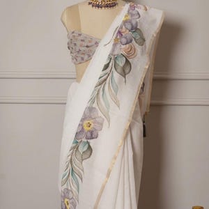 May include: A white saree featuring a floral pattern in purple, green, and gold. The saree is displayed on a mannequin, highlighting the hand-painted details. A matching blouse and necklace complete the outfit.