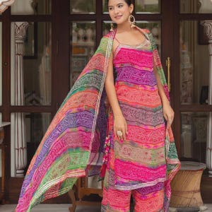 Puede incluir: Un vibrante conjunto indio de salwar kameez multicolor. El conjunto presenta una parte superior estampada en rosa, morado, verde y naranja, pantalones palazzo a juego y una dupatta fluida. El atuendo se complementa con joyas y sandalias doradas.