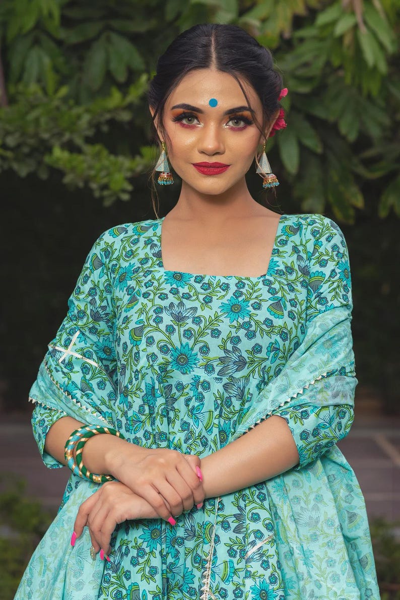 May include: A light blue square-neck kurta featuring a dark blue and green floral block print pattern, paired with a matching dupatta that has a silver scalloped trim along the edges, and green and gold bangles worn on the wrist.