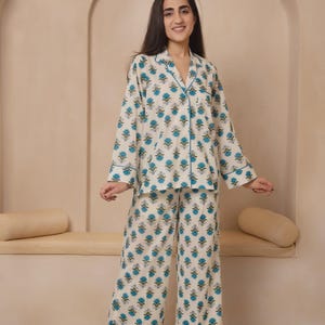 Könnte beinhalten: Zweiteiliger Pyjama in Creme mit einem türkisfarbenen Blumenmuster. Das langärmlige Oberteil und die weite Hose sind mit blauen Paspeln versehen. Das Pyjama-Set ist aus einem weichen, bequemen Material gefertigt.