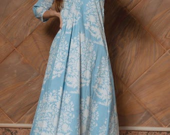 May include: A light blue dress with a white floral design. The dress features a V-neck and three-quarter sleeves. The model is wearing white trousers. The background is a brown, textured wall.