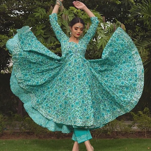 May include: A teal and blue floral print Anarkali dress with three-quarter sleeves and a flared skirt featuring silver metallic trim along the hem. The outfit includes matching teal cropped trousers with silver metallic horizontal stripes at the cuffs.