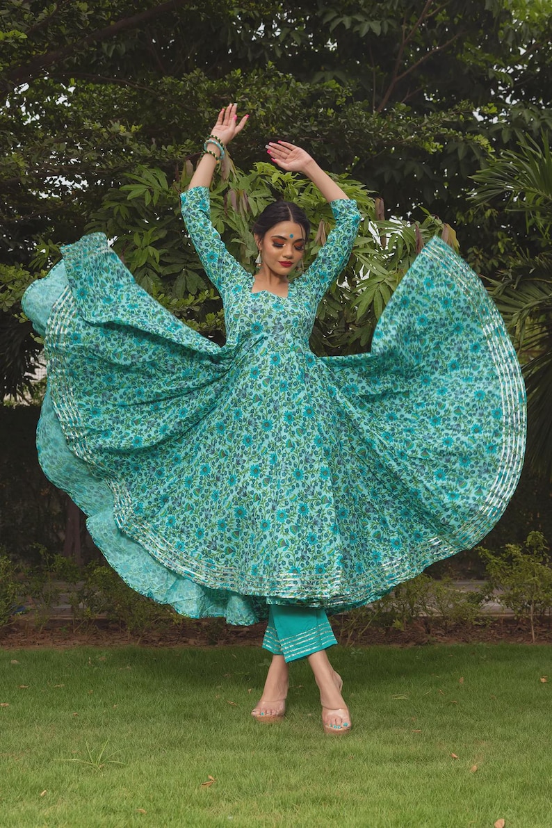 May include: A teal and blue floral print Anarkali dress with three-quarter sleeves and a flared skirt featuring silver metallic trim along the hem. The outfit includes matching teal cropped trousers with silver metallic horizontal stripes at the cuffs.