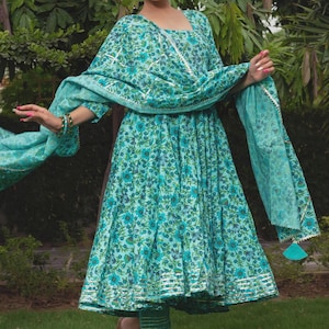May include: A turquoise floral print dress with a matching dupatta and trousers. The dress features a square neckline, three-quarter sleeves, and a flared skirt with silver metallic trim at the hem. The dupatta has a silver border and tassels.