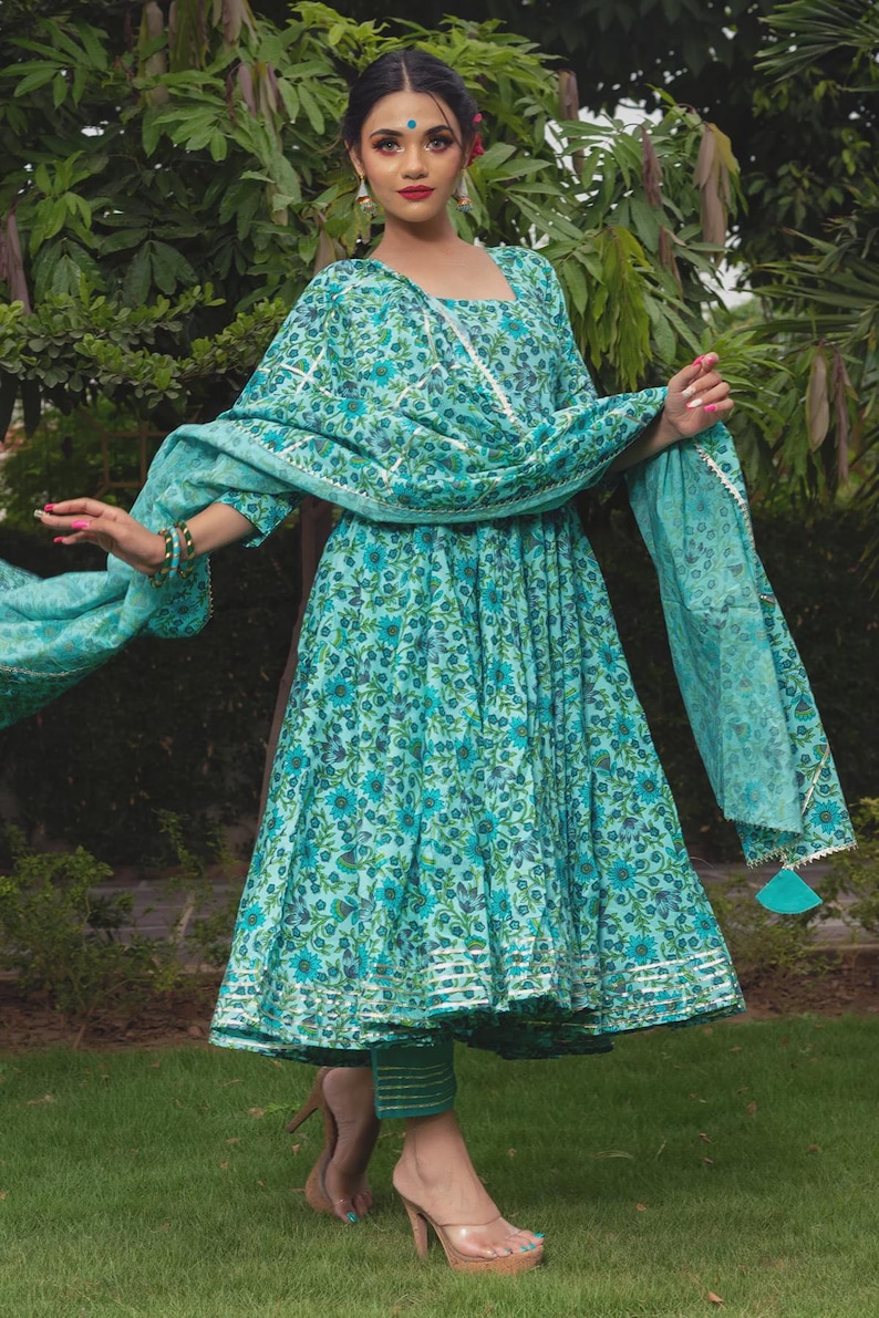 May include: A turquoise floral print dress with a matching dupatta and trousers. The dress features a square neckline, three-quarter sleeves, and a flared skirt with silver metallic trim at the hem. The dupatta has a silver border and tassels.