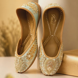 May include: A pair of light blue slip-on flats featuring intricate gold embroidery, small white pearls, and metallic thread detailing. The interior of the shoe is tan and displays the brand name Pomcha printed in white on the heel area.