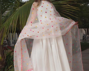 Puede incluir: Un vestido blanco largo hasta el suelo con cuerpo sin mangas y falda amplia. Un dupatta de organza blanca transparente se coloca sobre los hombros, con pequeños adornos de espejos circulares en amarillo y rosa, rematado con borlas rosas y amarillas.