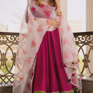 May include: A vibrant magenta-colored Indian salwar kameez ensemble. The outfit includes a long, flowing tunic, matching pants, and a sheer dupatta adorned with pink rose patterns. The dupatta has a gold trim and green tassels.