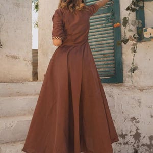May include: A long, flowing brown dress with short sleeves and a flared skirt. The dress features a high neckline and a subtle pattern on the sleeve cuffs. The person is wearing gold high heels. The background includes a white wall and a green shutter.