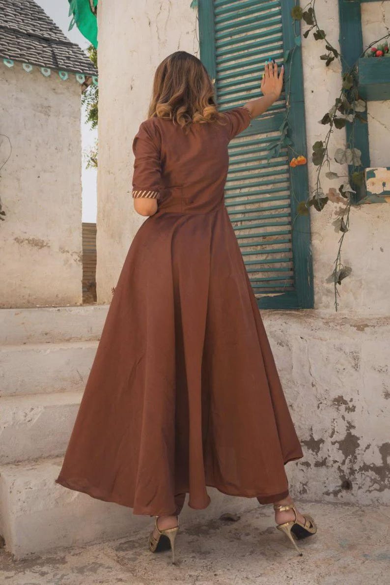 May include: A long, flowing brown dress with short sleeves and a flared skirt. The dress features a high neckline and a subtle pattern on the sleeve cuffs. The person is wearing gold high heels. The background includes a white wall and a green shutter.