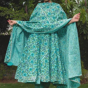 May include: A turquoise blue Anarkali dress featuring a dense floral pattern in darker blue and green tones. The dress includes a matching dupatta with silver trim and coordinating trousers with horizontal silver stripes at the hemline.