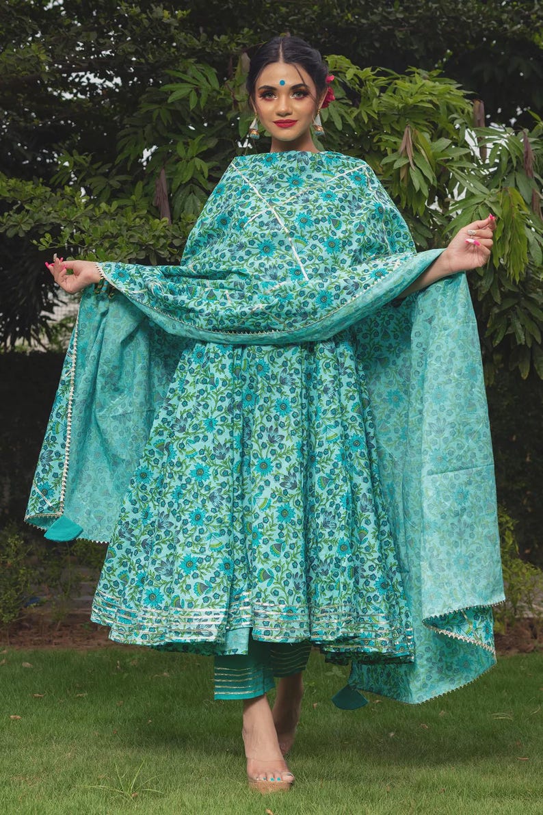 May include: A turquoise blue Anarkali dress featuring a dense floral pattern in darker blue and green tones. The dress includes a matching dupatta with silver trim and coordinating trousers with horizontal silver stripes at the hemline.