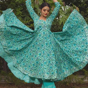 May include: A turquoise long-sleeved dress featuring a dense floral pattern in shades of blue and green, with a flared skirt and silver metallic trim along the hem. The dress is paired with matching turquoise cropped trousers with silver trim.