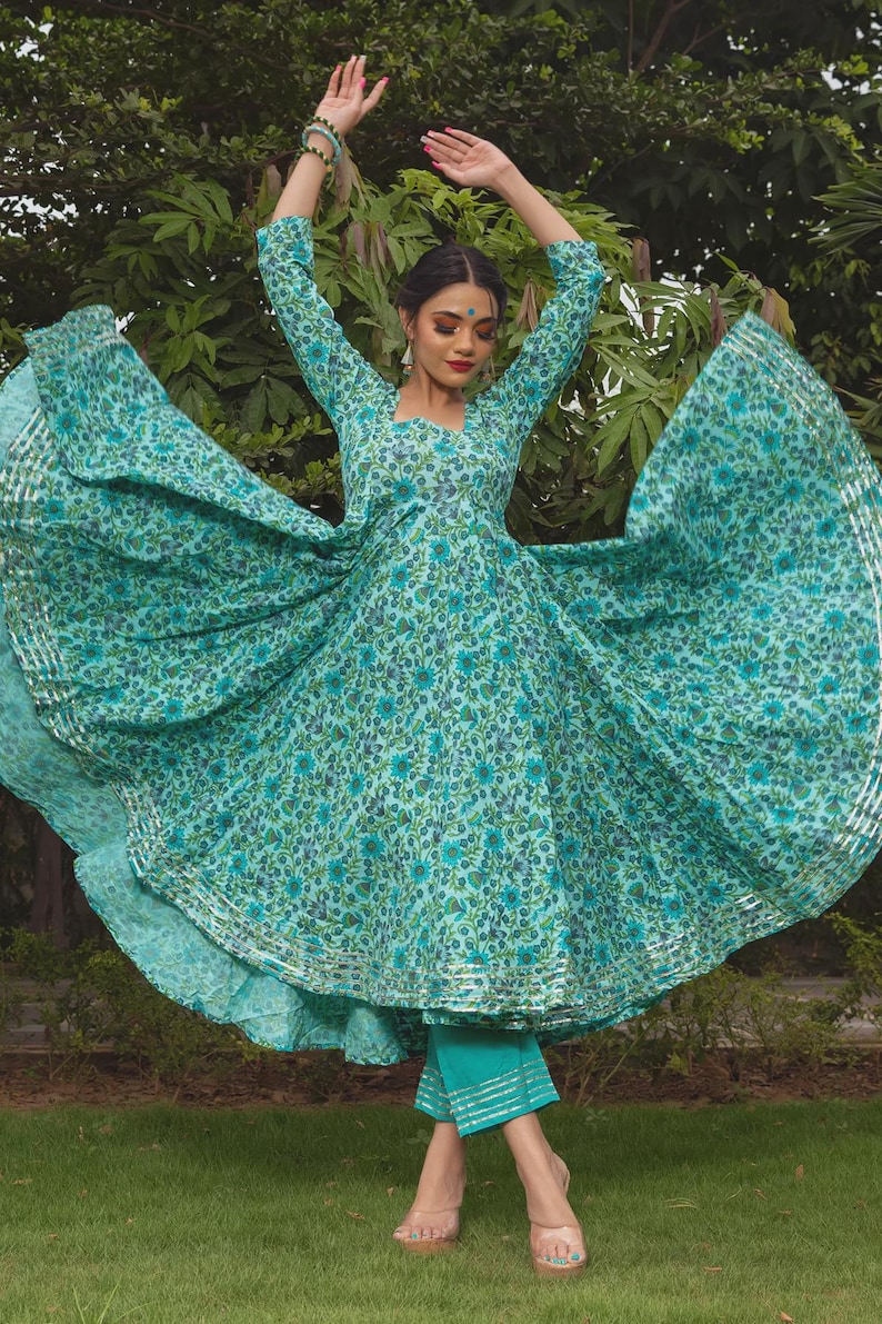 May include: A turquoise long-sleeved dress featuring a dense floral pattern in shades of blue and green, with a flared skirt and silver metallic trim along the hem. The dress is paired with matching turquoise cropped trousers with silver trim.