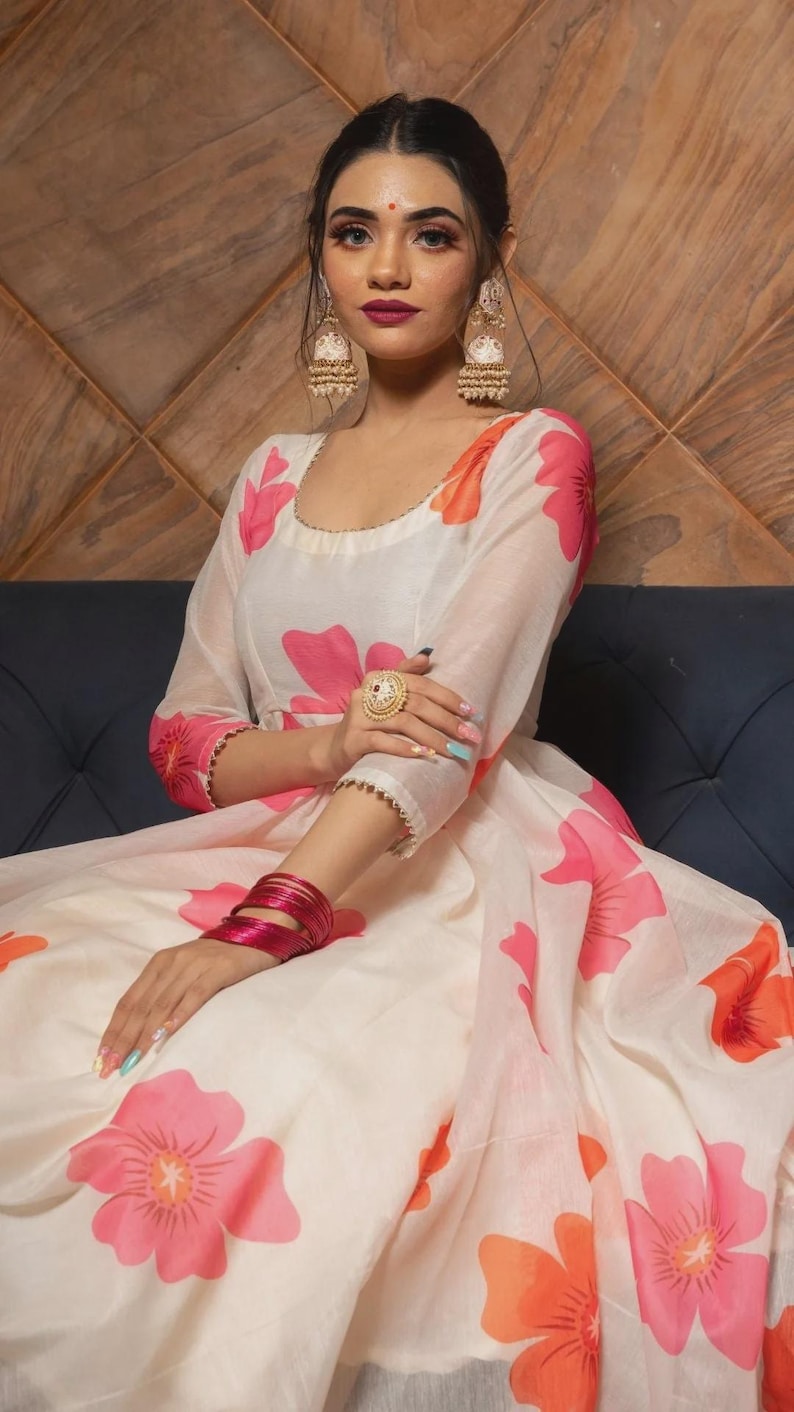 May include: A cream-colored floor-length dress featuring a vibrant floral pattern in shades of pink and orange. The garment has three-quarter length sleeves, a rounded neckline, and a full, flowing skirt. The fabric appears lightweight and semi-sheer.