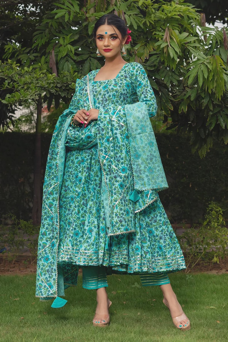 May include: A turquoise floral print long-sleeved dress with a matching dupatta and cropped trousers. The dress features a square neckline, a flared skirt with metallic trim, and a coordinating sheer scarf with decorative tassels and gold borders.