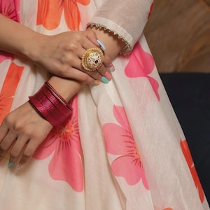 May include: A cream-colored dress featuring a large pink and orange floral print, three-quarter length sleeves with silver trim, a gold-toned ornate ring with a central dark stone, and a stack of thin, metallic red bangles on the wrist.