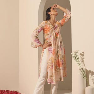 May include: A light peach-colored tunic with a floral pattern in shades of orange, purple, and pink. The tunic has a V-neck, long sleeves, and a flowing silhouette. Paired with white pants and embellished flats.