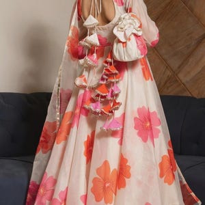 May include: A cream-colored A-line dress featuring a vibrant pink and orange floral print. The back includes a deep scoop neckline with tie closures and decorative hanging tassels in matching colors, paired with a small matching drawstring pouch.