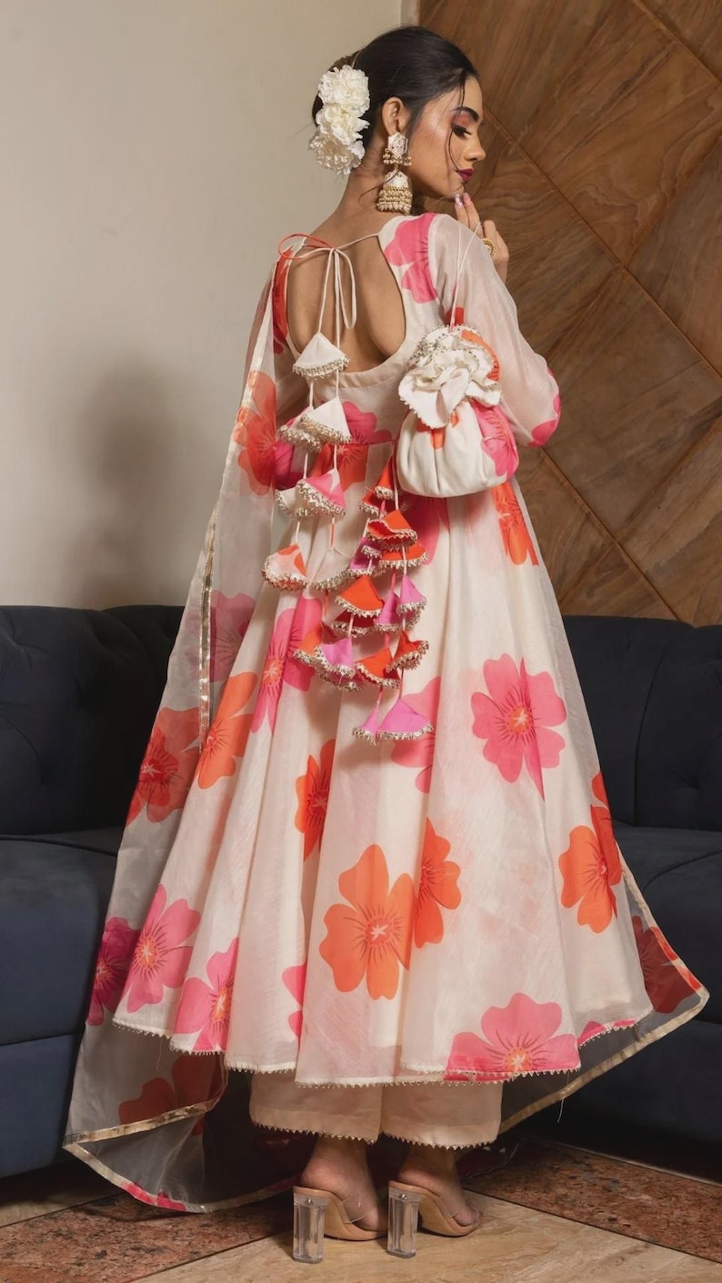 May include: A cream-colored A-line dress featuring a vibrant pink and orange floral print. The back includes a deep scoop neckline with tie closures and decorative hanging tassels in matching colors, paired with a small matching drawstring pouch.