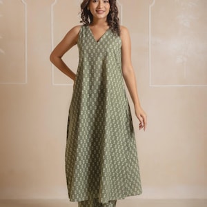 May include: A sleeveless, olive green, V-neck dress and matching pants set. The fabric features a repeating white floral pattern. The outfit is worn by a person. The background is a neutral beige with architectural details.