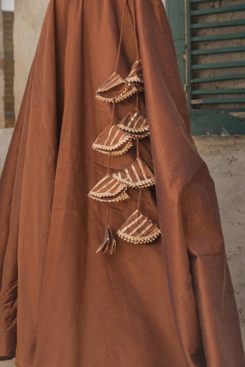 May include: A draped, brown fabric with decorative hanging tassels. The tassels are triangular, with a striped pattern in gold and brown, and trimmed with a white, scalloped edge. The fabric appears to be a heavy material.