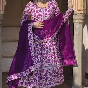 May include: A woman wearing a white and purple floral print salwar kameez with a matching dupatta. The outfit features a detailed floral pattern and is paired with matching pants. The dupatta is a solid purple color.