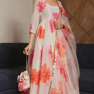 May include: A cream-colored Anarkali dress featuring a large pink and orange floral print, three-quarter sleeves, and a matching sheer dupatta with a metallic gold border. The outfit includes a small drawstring pouch with pink and orange tassels.