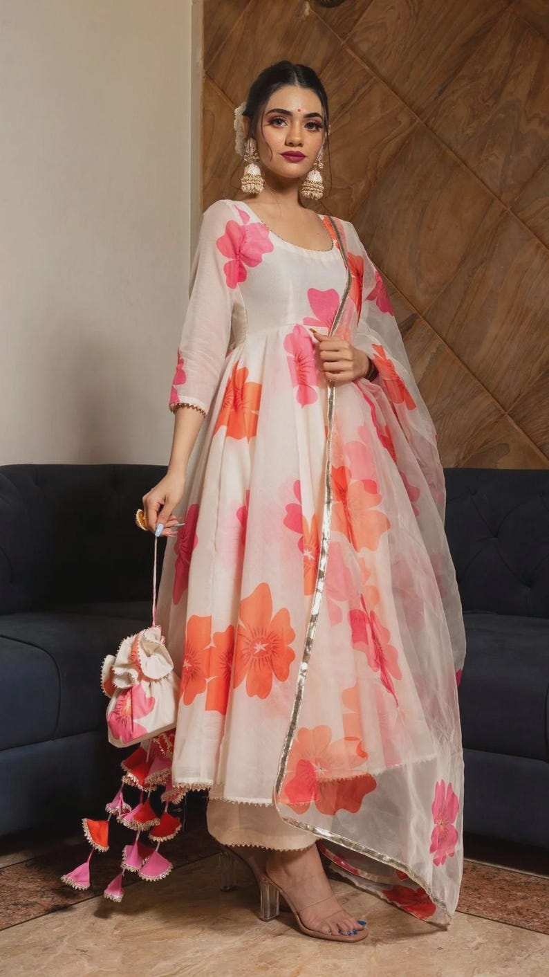 May include: A cream-colored Anarkali dress featuring a large pink and orange floral print, three-quarter sleeves, and a matching sheer dupatta with a metallic gold border. The outfit includes a small drawstring pouch with pink and orange tassels.