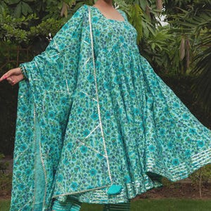 May include: A turquoise blue Anarkali dress featuring a dense floral pattern in darker shades of blue and green. The dress has a square neckline, long flared sleeves, and a matching dupatta with silver metallic trim and a tassel detail.
