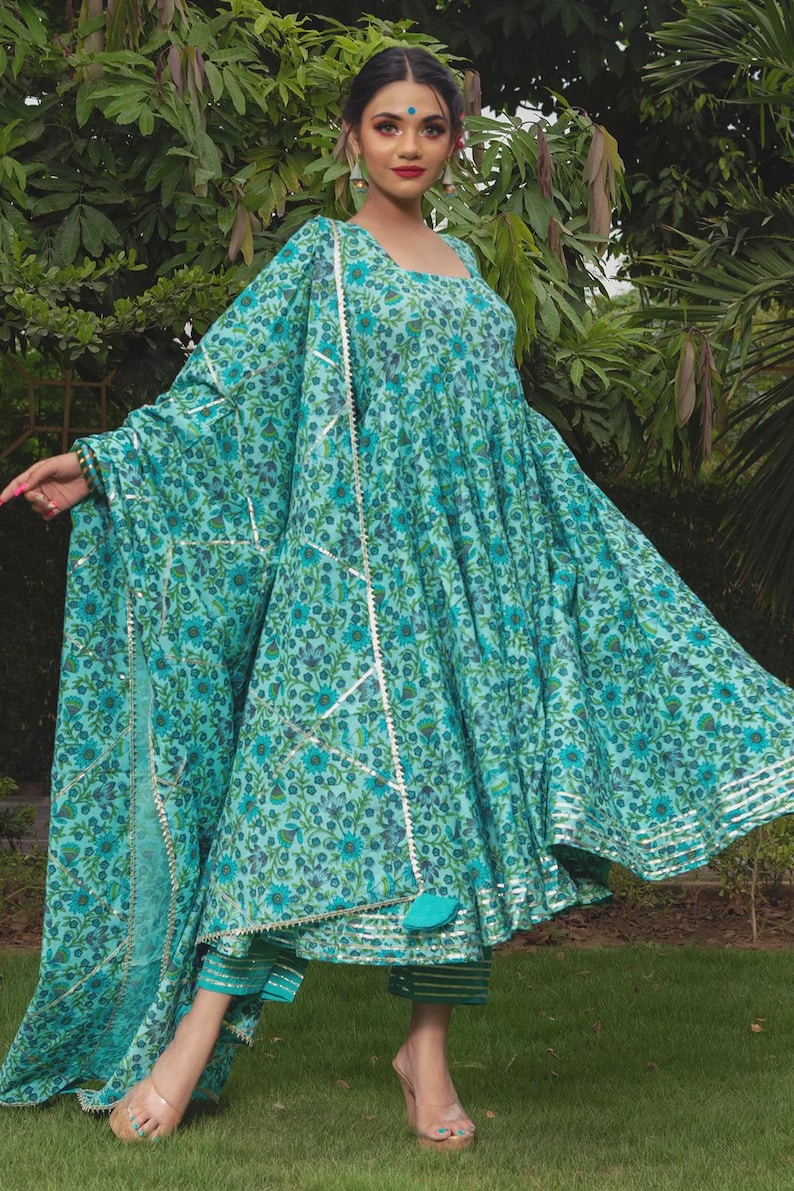 May include: A turquoise blue Anarkali dress featuring a dense floral pattern in darker shades of blue and green. The dress has a square neckline, long flared sleeves, and a matching dupatta with silver metallic trim and a tassel detail.