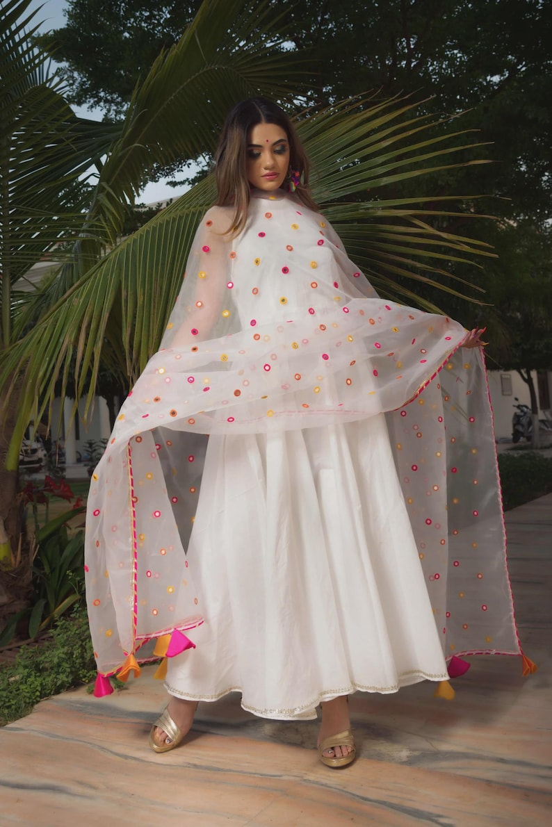 A white dress with a flowing skirt and a sheer, dotted shawl. The shawl is adorned with colorful circular accents and a pink trim. The outfit is completed with gold sandals and colorful tassels.