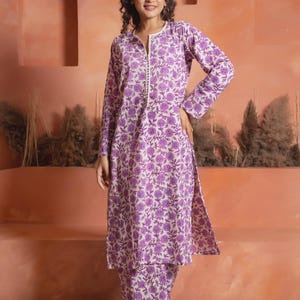 Op de afbeelding: Een tweedelige outfit met een tuniek met lange mouwen en een bijpassende broek. De set is versierd met een herhalend bloemenpatroon in paarse tinten op een crèmekleurige achtergrond. De tuniek heeft een split halslijn en een rechte snit.