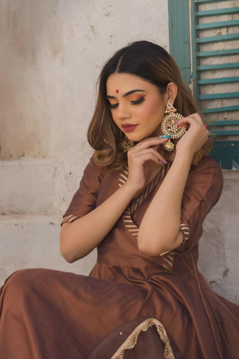 May include: A woman wearing a brown dress with gold trim and large, ornate gold earrings. The dress has short sleeves and a V-neck. The earrings are circular with intricate detailing. The woman has a red bindi on her forehead.