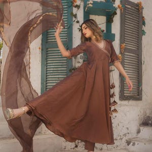 May include: A brown, flowing dress with a matching sheer scarf. The dress features a V-neckline and short sleeves with gold trim. The outfit is paired with gold sandals. The background includes a white wall and shuttered windows.